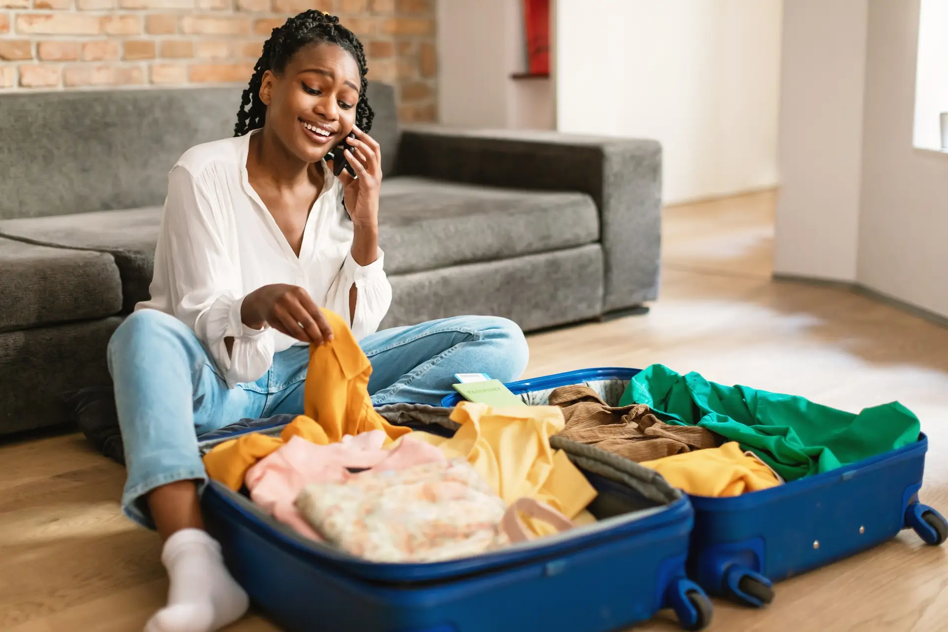 Happy black woman having phone call while packing clothes into travel bag, preparing for vacation